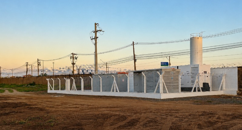 Subestação de entrada de energia cercada, com cabines, gerador e rede aérea ao fundo